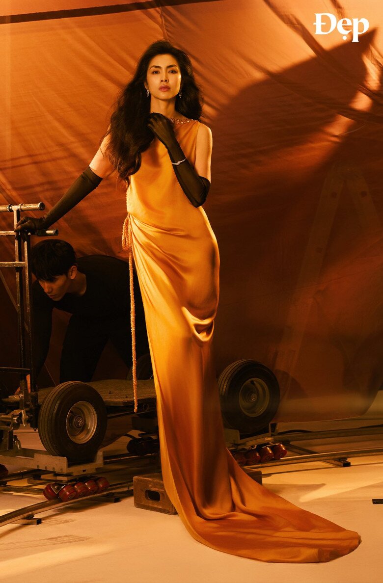 Trong khung hình thứ hai, Tăng Thanh Hà kiêu sa trong thiết kế đầm lụa vàng đồng có độ bóng nhẹ, ôm sát cơ thể và đổ dài chạm sàn. Chất liệu satin mềm mại bắt sáng tinh tế, tôn lên làn da mịn màng và vóc dáng thanh thoát của cô. Phụ kiện găng tay đen dài quá khuỷu tay mang đến điểm nhấn sang trọng, vừa cổ điển vừa quyền lực.