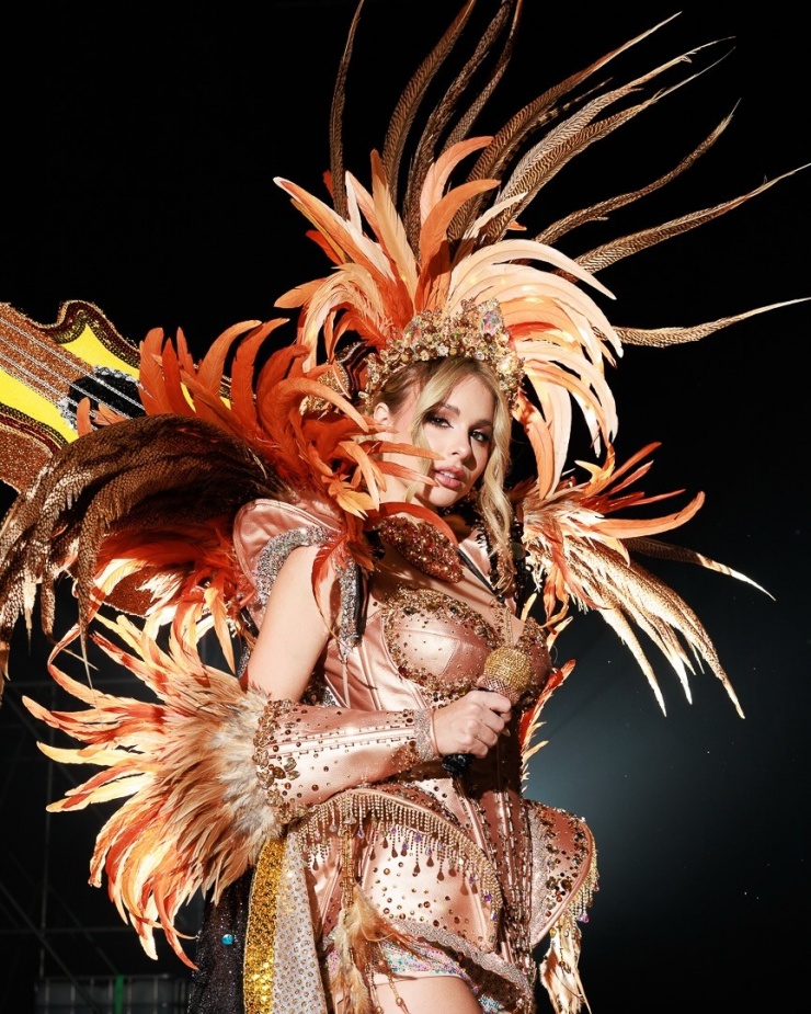 Trang phục dân tộc đa sắc màu của thí sinh Miss Universe 9 Miss Universe Argentina 2025 Aldana Masset xuất hiện trong bộ trang phục được thiết kế theo phong cách nghệ sĩ trình diễn lễ hội Carnival. Cô 25 tuổi, biết chơi ba loại nhạc cụ. Năm 2022, cô giữ vai trò giọng hát chính của ban nhạc Agapornis.