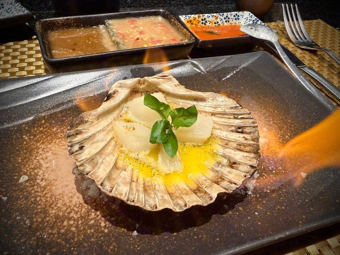 Khoảnh khắc đời thường nhưng ngọt ngào ấy nhanh chóng nhận được sự quan tâm của người hâm mộ. Đặc biệt, trong loạt món được gọi, món sò điệp lại bất ngờ trở thành tâm điểm chú ý. Theo hình ảnh được Trấn Thành chia sẻ, món sò điệp áp chảo/nướng bơ được phục vụ theo phong cách cao cấp. Món ăn gây ấn tượng ngay từ hình thức khi sò được đặt trên chính vỏ sò điệp, phía dưới là lớp bơ tan chảy vàng óng. Khi mang ra bàn, món ăn vẫn còn lửa cháy nhẹ, vừa giữ nhiệt vừa tạo hiệu ứng thị giác đầy cuốn hút, đúng chuẩn phong cách ẩm thực fine dining.